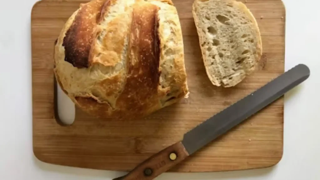 Freshly baked gluten free sourdough loaf on cooling rack with slice cut open showing airy crumb, styled with bread knife, linen towel, and olive oil dish