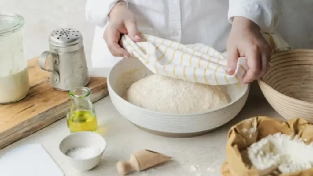 Gluten free sourdough dough proofing in loaf pan covered with kitchen towel, rustic baking setup with starter jar and flour